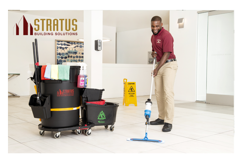 A Smiling Uniformed Stratus Cleaner with a Trash Can and Mop Bucket on Wheels with Cloths on it Next to Him Mops a Tile Floor